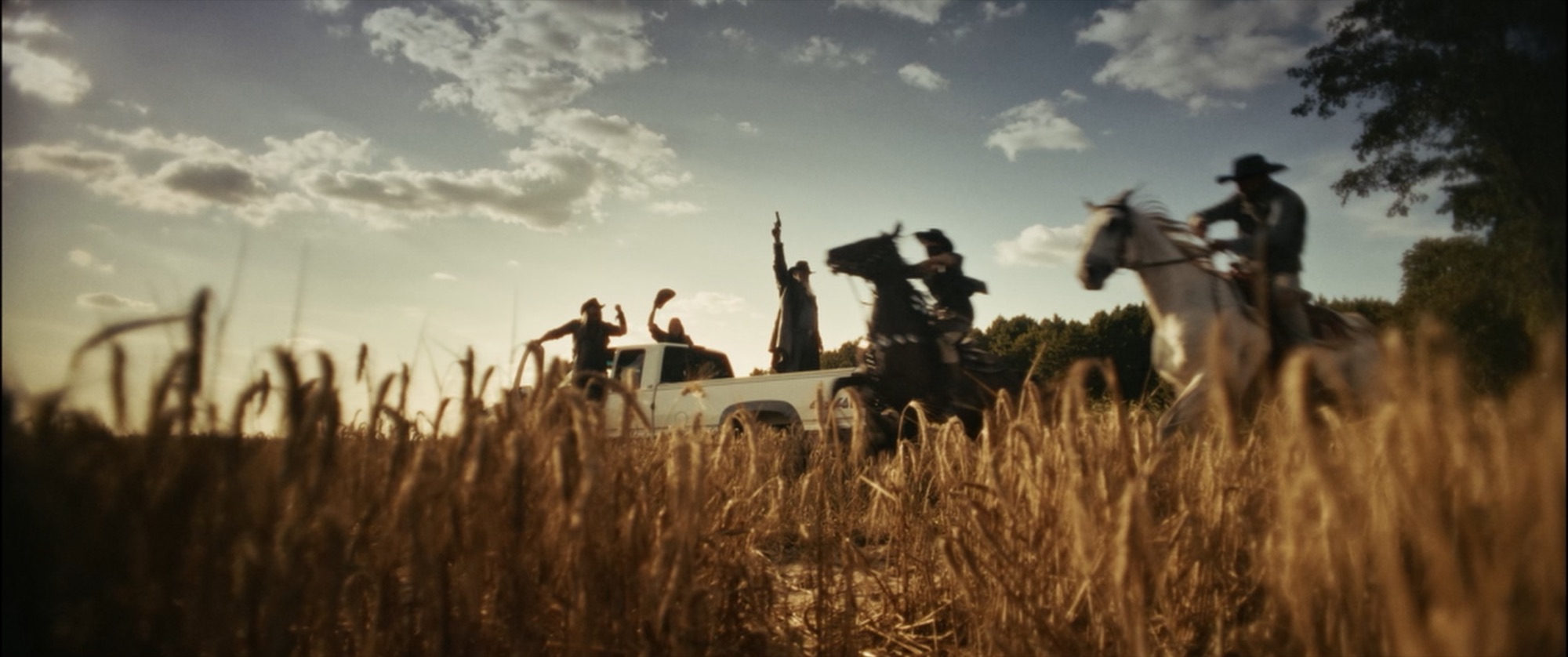 Cowboys on horseback and a pickup in a wheat field, seen low through the grain, one arm raised in golden light - Amon Amarth